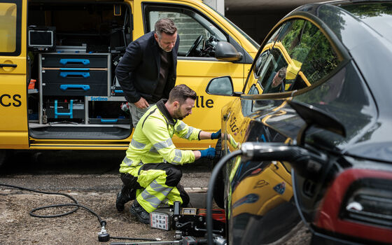 Une personne vêtue d'un gilet de sécurité travaille sur la roue d'une voiture électrique noire. À côté, une autre personne se tient devant un véhicule de service jaune dont le coffre est ouvert et qui contient des outils.